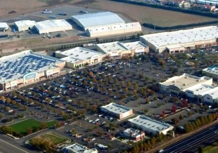 A shopping center built by Hilbers. It is an aerial view with a parking lot closest and a group of light-colored buildings with solar panels farther back, and the sky is blue.