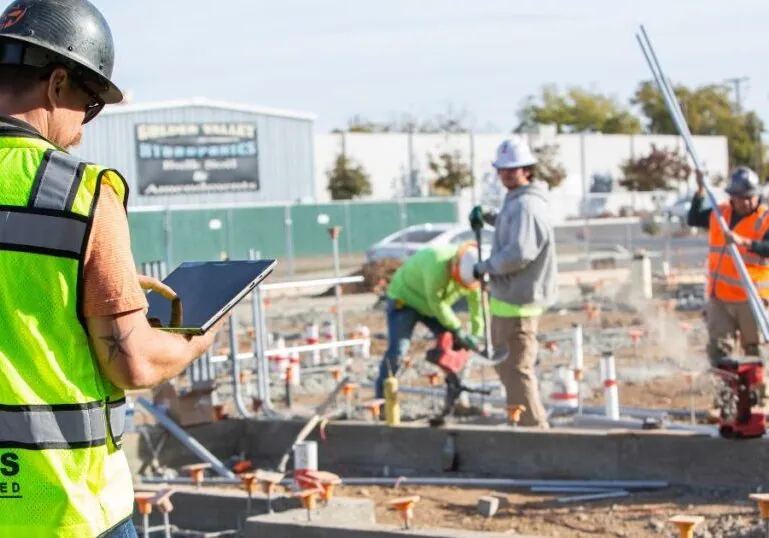 A man in a Hilbers high-vis vest points at a tablet in front of a construction site. Three workers are working on the site, holding various materials.