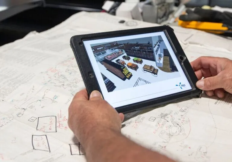 Two hands hold a tablet over architectural plans. The tablet displays a computer model of the finished building.