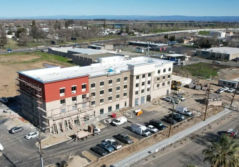 An aerial view of a large building with scaffolding on the side. Cars are parked in front and there is a dirt lot behind it.